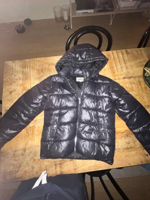 Svart vinterjacka  från ONLY med huva - Säljer en svart pufferjacka från ONLY med huva och dragkedja framtill. Jackan har två fickor med knappstängning och ett märke på ena ärmen. Perfekt för kalla dagar och har en snygg, glansig finish. Moncler liknande. 