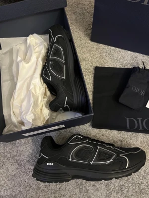 Svarta Dior B30 sneakers - Säljer ett par svarta Dior B30 sneakers med coola vita detaljer och logga på sidan. Skorna har en chunky platt sula, rund tå och snörning. Materialet är en mix av mesh och syntet med snygga kontrastlinjer som ger en modern vibe. Perfekta för dig som gillar exklusiva märken och streetstyle.