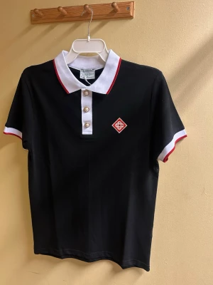 Svart polotopp från Casablanca - Svart polotopp från Casablanca med vit krage och röda detaljer. Toppen har korta ärmar med vit och röd kant, tre pärlemorknappar framtill och ett broderat rött emblem på bröstet. Materialet är mjuk bomull som känns skönt mot huden.