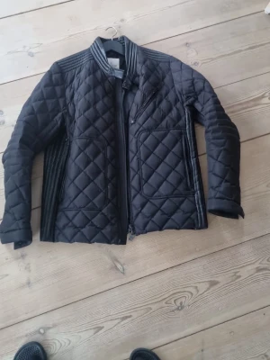 Svart quiltad dunjacka från Moncler - Snygg svart dunjacka från Moncler med quiltat rutmönster och coola detaljer på axlar och ärmar. Jackan har ståkrage, dragkedja framtill och praktiska fickor med zip. Perfekt för dig som vill ha en stilren och varm jacka med exklusiv känsla.skriv om intresse hör av dig om du har fårger 