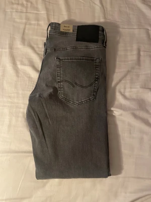 Grå jeans från Jack & Jones - Snygga grå jeans från Jack & Jones med klassisk femficksdesign och svarta detaljer på bakfickorna. Jeansen har en rak passform och är tillverkade i mjukt denim-material som sitter skönt hela dagen. Perfekt för dig som gillar stilrena och enkla jeans.
