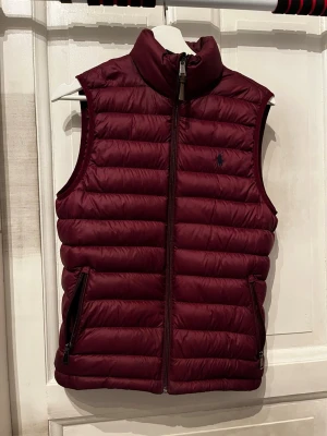 Vinröd dunväst från Polo Ralph Lauren - Snygg vinröd dunväst från Polo Ralph Lauren med quiltad design och mörkblå logga på bröstet. Västen har två dragkedjeförsedda fickor och hög krage. Perfekt att slänga över en hoodie för en chill och stilren look.