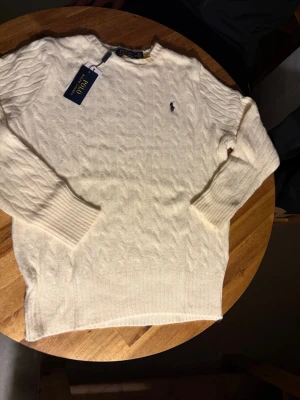 Vit kabelstickad tröja Polo Ralph Lauren - Vit kabelstickad tröja från Polo Ralph Lauren i storlek M. Tröjan har långärmade ribbade muddar och klassisk rund hals. Den ikoniska Polo-loggan är broderad på bröstet. Perfekt för dig som gillar stilren och tidlös design.