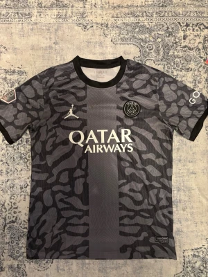 PSG x Jordan svart leopard fotbollströja - Säljer en PSG x Jordan fotbollströja i svart och grått med leopardmönster. Tröjan har Qatar Airways-tryck på bröstet, GOAT på ärmen och Ligue 1-märke. Rund hals och snyggt PSG-emblem på bröstet. Perfekt för dig som älskar streetwear och fotboll.