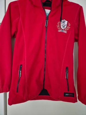 Fleecejacka - Röd fleecejacka från Mywear Active med huva, broderad RC Royal 1884 Club-logga på bröstet och två fickor med dragkedja. Jackan har hel dragkedja framtill och elastiska muddar vid ärmslut. Perfekt för dig som gillar sportig och bekväm stil. Finns några fläckar.