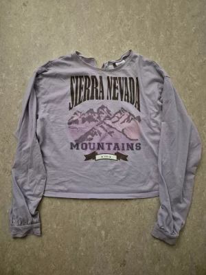 Tröja - Ljuslila croppad tröja från H&M med tryck 'Sierra Nevada Mountains' och bergmotiv i lila toner. Tröjan har lång ärm, rund halsringning med liten v-formad öppning och är gjord i mjuk bomull. Perfekt för dig som gillar chill och avslappnad stil.