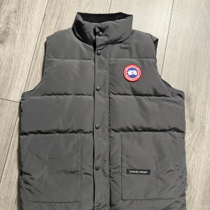 Svart dunväst från Canada Goose - Svart dunväst från Canada Goose med hög krage, två stora fickor framtill och klassisk logotyp på bröstet. Västen är quiltad och har tryckknappar längs hela framsidan. Perfekt för kyliga dagar när du vill hålla stilen.