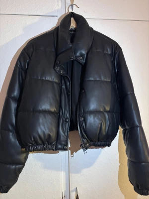 Svart skinnpufferjacka  - Säljer en svart pufferjacka från Zara i storlek S. Jackan har en croppad passform, quiltad design och är tillverkad i syntetmaterial med skinnlook. Den har dragkedja framtill, tryckknappar vid kragen och elastisk nederkant för en cool siluett.