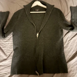 Mörkgrå kofta med dragkedja från H&M - Snygg mörkgrå kofta från H&M med hel dragkedja framtill och hög krage. Tröjan är långärmad. Materialet är mjukt och känns som en blandning av bomull och syntet. Perfekt för kyliga dagar. Ny pris är 449.