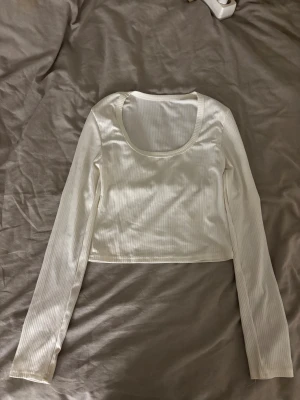 Vit ribbad croppad långärmad topp - Säljer en vit croppad topp med långa ärmar och ribbad struktur. Toppen har en rundad, lite djupare halsringning och är tillverkad i ett stretchigt material som sitter snyggt på kroppen. Perfekt för dig som gillar stilrena och enkla plagg.