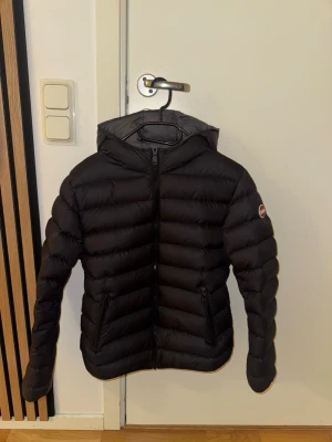 Svart dunjacka från Colmar med huva - Säljer en svart dunjacka från Colmar med huva och två sidofickor med dragkedja. Jackan har ett stilrent, quiltat mönster och är tillverkad i polyester med dunfyllning. Perfekt för kalla dagar och har en snygg, normal passform. Ikonisk Colmar-logga på ärmen.