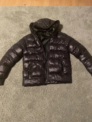 Svart pufferjacka SHEIN  - Svart pufferjacka från Fashion Man i storlek S. Jackan har en glansig yta, huva, två fickor med dragkedja och ribbade muddar vid ärmslut. Materialet är syntet och jackan stängs med en rejäl dragkedja framtill. Perfekt för kalla dagar.