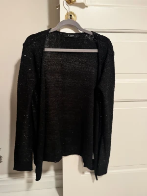 Svart glittrig kofta från Vila - Svart öppen kofta från Vila med glittriga paljettdetaljer. Långärmad och stickad i ett lätt material som ger en snygg och luftig känsla. Perfekt för utgång eller nyår 🍾🎊