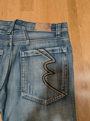 Blå Diesel jeans med broderade fickor - Snygga blå jeans från Diesel med klassisk tvättad look och raka ben. Baksidan har broderade fickor med zigzag-mönster i beige och mörkblått. Framtill finns Diesel Industry-logga på myntfickan. Perfekt för dig som gillar street och casual.