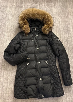 Svart lång pufferjacka med päls - Svart pufferjacka från Rockandblue Collection med quiltade detaljer och päls på huvan. Jackan har flera dragkedjor, knappar framtill och en patch på ärmen. Perfekt för kalla dagar med sin långa passform och snygga look.