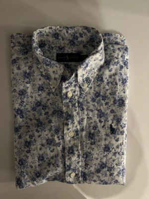 Blommig skjorta från Ralph Lauren - Vit skjorta från Ralph Lauren med blått blommigt mönster och klassisk krage. Skjortan har knappar framtill och broderad logga på bröstet. Tillverkad i bomull, perfekt för dig som gillar stilrena men ändå unika plagg. DEFEKT sista bilden