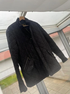 Svart quiltad jacka från Barbour - Svart quiltad jacka från Barbour med klassisk krage och tryckknappar framtill. Jackan har två fickor med lock och dragkedja, samt ett stilrent rutmönster över hela ytan. Perfekt för dig som gillar tidlös och clean stil.