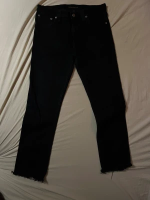 Svarta Jeans från Nudie Jeans Co W30 L32 - Svarta jeans från Nudie Jeans Co i klassisk femficksmodell. Jeansen har råa, avklippta benslut med fransar för en edgy vibe. Passformen är rak och materialet är slitstarkt denim. Perfekt för dig som gillar minimalistisk stil med en twist. Hör av dig vid frågor