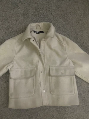 Beige kappa från Zara - Snygg beige kappa från Zara med klassisk krage och stora fickor framtill. Jackan har tryckknappar och är tillverkad i ett mjukt material. Perfekt för lager-på-lager och har en clean, stilren look som funkar till många outfits. 