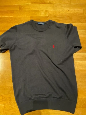Svart/kortärmad tröja Polo Ralph Lauren - Svart kortärmad tröja från Polo Ralph Lauren med klassisk rund hals och röd broderad logga på bröstet. Tröjan är i mjuk bomull och har ribbade muddar vid hals och nederkant. Perfekt för dig som gillar stilrena och tidlösa plagg.