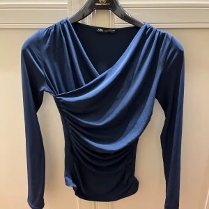 Mörkblå omlottopp från Zara - Säljer en mörkblå omlottopp från Zara med snygg drapering och v-ringad hals. I storlek S. Använd ett fåtal gånger så fortfarande bra skick 