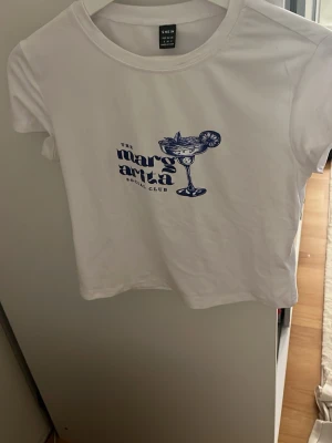 Vit t-shirt med blått tryck från SHEIN - Snygg vit t-shirt från SHEIN i storlek S med ett blått tryck på bröstet som föreställer en margarita och texten 'The Margarita Social Club'. T-shirten har rund hals och korta ärmar. Den är använd ett fåtal gånger men då har de bara varit hemma