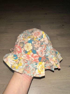 Blommig solhatt med volangkant Flerfärgad solhatt - Söt solhatt med blommigt mönster i pastellfärger som rosa, gul, blå och beige. Hatten har en bred volangkant runt hela och är tillverkad i mjuk bomull. Perfekt för att ge din look en färgglad och lekfull touch under soliga dagar.Söt solhatt med ett blommigt mönster och en bred brätte som ger bra skydd mot solen. För 3 mån +