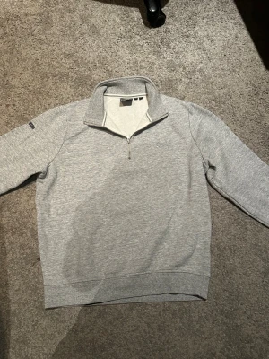 Grå half zip tröja från Superdry - Snygg grå långärmad tröja från Superdry med halv dragkedja och broderad logga på bröstet. Tröjan har en mjuk insida och ribbade muddar vid ärmslut och nederkant. Perfekt för chill dagar och enkel att matcha med jeans eller joggers.