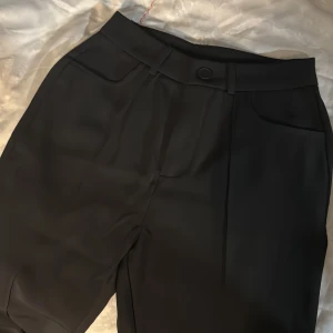 Svarta kostymbyxor med knapp - Snygga svarta bootcut kostymbyxor med hög midja. Etiketten på byxorna sitter kvar och är helt oanvända. Byxorna har framfickor, bälteshällor och stängs med knapp och dragkedja. Materialet är mjukt och består av 94% polyester och 6% elastan för extra komfort.