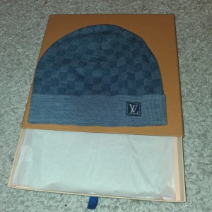 Blå rutig mössa från Louis Vuitton - Snygg blå mössa från Louis Vuitton med diskret rutigt mönster och uppvikt kant. LV-logga framtill och tillverkad i 100% ull för extra värme. Perfekt accessoar för dig som vill ha en lyxig och stilren look.