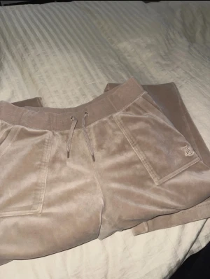 Beiga velourbyxor Juicy Couture M - Säljer ett par beiga mjukisbyxor från Juicy Couture i mjuk velour. Byxorna har snörning i midjan, fickor fram och bak med knappdetalj samt broderad logga på ena fickan. Perfekta för en chill och trendig look. Saknar silver detaljerna på snörningen!