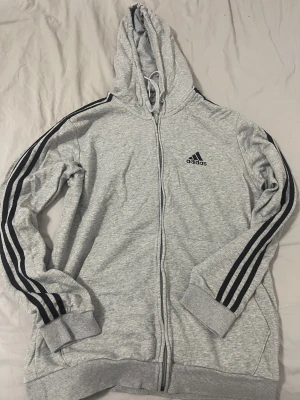 Grå Adidas hoodie med svarta ränder - Säljer en grå hoodie från Adidas i storlek L. Tröjan har klassiska svarta ränder längs ärmarna, dragkedja framtill och huva med snörning. Adidas-logga på bröstet och ribbade muddar vid ärmslut och nederkant. Perfekt för chill eller träning.