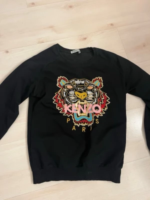 Svart sweatshirt med tiger från Kenzo - Svart sweatshirt från Kenzo med ikonisk broderad tiger i flera färger på bröstet och texten 'KENZO PARIS' under. Tröjan har rund hals, långa ärmar och ribbade muddar. Perfekt statement-plagg för dig som gillar att sticka ut.