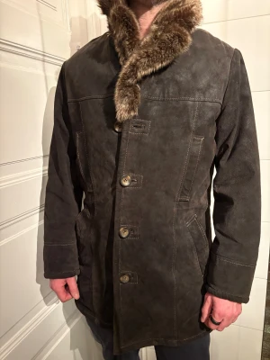 Brun mocka rock med pälsdetalj JOFAMA stl.M - Säljer en brun rock i mocka med bred, fluffig päls runt kragen. Jackan har synliga sömmar, knäppning med stora knappar och två fickor framtill. Supermjukt fluffoder innuti. Perfekt för dig som vill sticka ut med en klassisk men ändå unik stil.Märke; Jofama. Några lagningar vid 2 knappar. Inget som syns. Sista bilden är lagningar fotade.