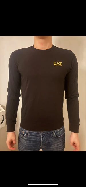 Svart långärmad tröja EA7 - Svart långärmad tröja från EA7 Emporio Armani med gul logga på bröstet och gul randdetalj i nacken. Tröjan har rund hals och är tillverkad i mjuk bomull som sitter snyggt och bekvämt. Perfekt för dig som gillar stilrena och sportiga plagg.