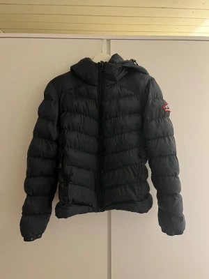 Svart pufferjacka Superdry Fuji - Svart pufferjacka från Superdry Fuji Series med huva och dragkedja framtill. Jackan har quiltad design, två sidofickor med dragkedja och en snygg patch på ärmen. Perfekt för kalla dagar och har en modern, sportig look. Köpt på superdry för 999kr.