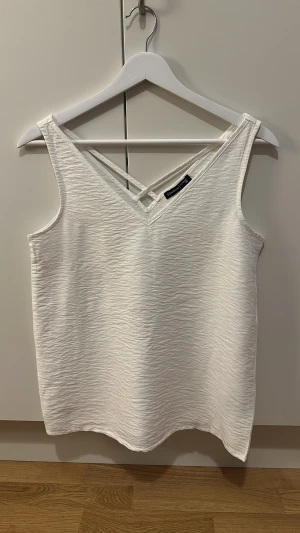 Vit linnetopp med v-ringning LC Waikiki - Säljer en fräsch vit linnetopp från LC Waikiki i storlek 36. Toppen har en snygg v-ringning med dubbla band framtill och ärmlös design. Materialet är lätt strukturerat och känns mjukt mot huden, perfekt för varma dagar. Enkel och stilren look.