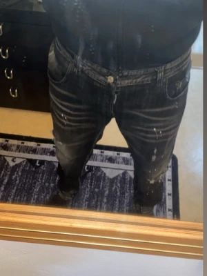 Svarta Dsquared2 jeans med detaljer - Säljer ett par svarta jeans från Dsquared2 med coola slitningar och färgstänk i rött och vitt. Jeansen har en straight fit och klassisk femficksdesign. Materialet är jeans och de har en unik patch med Dsquared2-logga på bakfickan.
