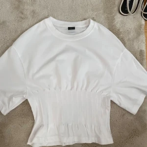 Vit topp från Gina Tricot - Säljer en vit croppad topp från Gina Tricot i storlek S. Toppen har korta ärmar och ett ribbat parti nedtill som ger en snygg form. Materialet är mjuk bomull och modellen är loose med rund hals. Perfekt för dig som gillar stilrena och enkla plagg.