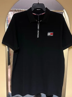 Svart polotopp från Tommy Hilfiger - Svart polotopp från Tommy Hilfiger med klassisk krage och kort ärm. Toppen har en dragkedja framtill med logotyp och ett broderat märke på bröstet i rött, vitt och blått. Materialet är mjuk bomull som känns skönt mot huden. Perfekt för en clean och stilren look.