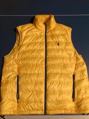 Gul dunväst från Polo Ralph Lauren - Snygg gul dunväst från Polo Ralph Lauren med klassisk broderad logga på bröstet. Västen har ståkrage, två fickor med dragkedja och quiltad design. Perfekt för lager-på-lager och ger en cool vibe till din outfit.