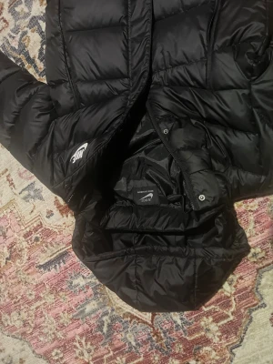 Svart lång dunjacka från Nike - Säljer en svart lång dunjacka från Nike med huva och vita logotypdetaljer på bröstet och ärmen. Jackan har dragkedja, knappar och två stora fickor framtill. Perfekt för kalla dagar med sin quiltade design och stilrena look.