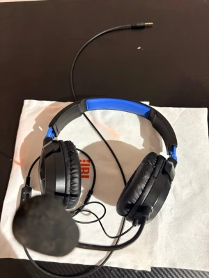 Turtle Beach Recon 70 Gaming Headset - Turtle Beach Recon 70 gaming headset med kabel och mikrofon. Over-ear design, blå/svart färg. Synligt slitage på öronkuddarna, men fungerar bra för spel och kommunikation. 3,5 mm anslutning, kompatibel med flera plattformar.