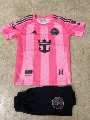 Inter Miami Messi fotbollströja set - Säljer ett Inter Miami fotbollströja och shorts set från Adidas, med Messi och nummer 10 på ryggen. Tröjan är rosa med svarta detaljer och ränder på axlarna, samt klubbmärke och sponsortryck. Shortsen är svarta med rosa klubbmärke. Perfekt för dig som älskar fotboll och Messi.