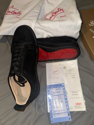 Christian Louboutin svarta sneakers - Svarta sneakers från Christian Louboutin med lyxig mocka och signaturröd sula. Snygg broderad logga på hälen och klassisk snörning framtill. Insidan är ljusbeige och skon har en rund tå. Perfekt för dig som vill sticka ut med exklusiva detaljer.