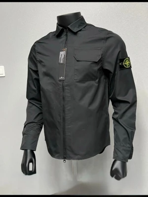 Svart overshirt från Stone Island - Svart overshirt från Stone Island med klassisk krage, bröstficka med lock och ikonisk patch på vänster ärm. Jackan har hel dragkedja framtill och är tillverkad i ett lätt syntetmaterial. Perfekt för dig som gillar clean och stilren streetwear.