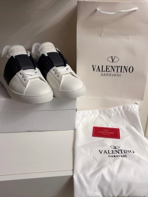 Valentino Garavani Open sneakers vit/svart - Säljer ett par Valentino Garavani Open sneakers i vitt skinn med bred svart rem över mitten. Klassisk rund tå, platt sula och coola nitar baktill på hälen. Skorna har vita snören och kommer med originalpåse och box. Perfekt för dig som gillar lyxiga detaljer.