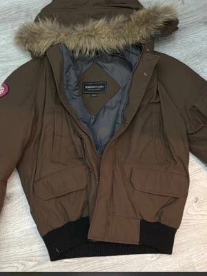 Brun bomberjacka med fuskpäls - Snygg brun bomberjacka från Noble Bird Classics med huva och fuskpäls. Jackan har flera fickor framtill, ribbade muddar och dragkedja. Perfekt för dig som vill ha en cool och varm jacka med streetkänsla.