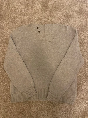 Ralph Lauren Kofta/Tröja - Ralph Lauren stickad tröja – stl L  Klassisk och stilren stickad tröja från Ralph Lauren i en neutral grå nyans. Modellen har två dekorativa knappar som man kan knäppa på 2 olika sätt vilket ger ett lite mer unik och dressad look. Perfekt både till vardags och lite finare tillfällen.  🔹 Storlek: Large 🔹 Färg: Grå/Vit 🔹 Skick: Mycket fint skick, nästan nyskick. – inga synliga defekter, hål eller fläckar. 🔹 Material: Kontrollera gärna tvättlappen (ej synlig på bilden). 🔹 Nypris: Ca 1700-2000kr ber