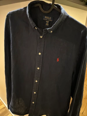 Mörkblå skjorta från Polo Ralph Lauren - Snygg mörkblå skjorta från Polo Ralph Lauren i storlek XL i barn så passar XS/S också . Skjortan har klassisk krage, knappar framtill och den ikoniska röda Polo-loggan broderad på bröstet. Tillverkad i mjukt linne, perfekt för en chill och stilren look. Priset är inte hugget i sten!
